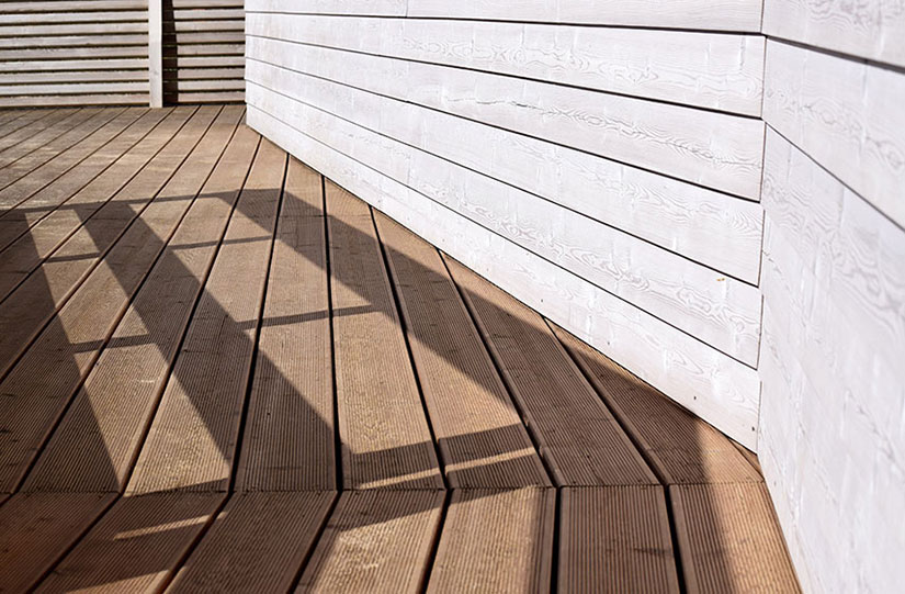 A deck with brown deck boards beside a white wall.
